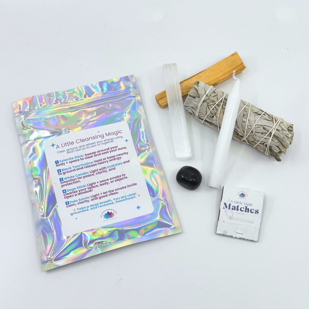 A collection of items for an energy cleansing kit including a selenite stick, black tourmaline, Palo Santo stick, sage bundle, white candle with matches, and packaging with a reflective surface.