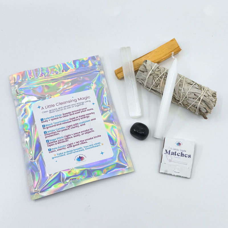 A collection of items for an energy cleansing kit including a selenite stick, black tourmaline, Palo Santo stick, sage bundle, white candle with matches, and packaging with a reflective surface.