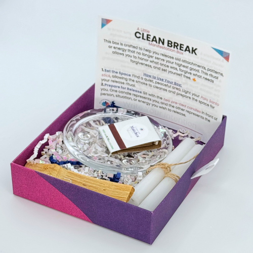A Clean Break Manifestation Box opened to reveal a glass holder with candles, a palo santo stick, and a piece of cloth with a twine knot inside. The box is labeled with instructions and a list of included items.